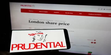 A smartphone displays the Prudential logo in front of a computer screen showing the Prudential website with the "London share price" section open. The setup highlights Prudential's branding and a segment of their online financial information, emphasizing their position within the FTSE 100. | FintechZoom