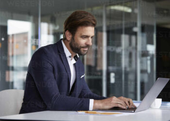 A man with a beard, wearing a navy suit and white shirt, is sitting at a table in a modern office, working on his laptop. The large glass windows in the background reflect his focus on quality management. | FintechZoom
