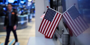 A small American flag is mounted on a desk with its reflection visible on a nearby surface. In the background, there is a person walking across the trading floor of a stock exchange, with monitors and electronic boards displaying information from the stock market today. | FintechZoom