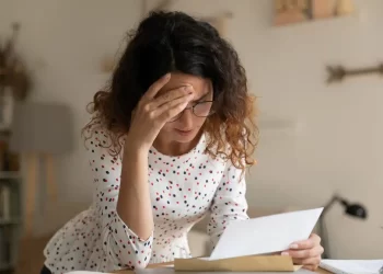 A woman with curly hair and glasses is leaning over a desk, holding her forehead with one hand and reading a piece of paper about unexpected expenses with the other. She appears to be stressed or worried. She is wearing a white shirt with red and black polka dots. | FintechZoom