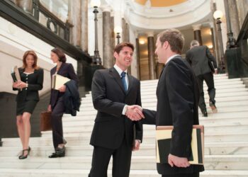 Two businessmen in suits shake hands on a grand staircase in a large, elegant building with marble columns. Three additional professionals, including a workers compensation attorney, walk up and down the stairs in the background, all holding folders or briefcases. | FintechZoom