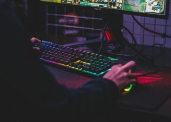 A person is playing a computer game, seen from the side. They are using a colorful RGB backlit keyboard and mouse, with a gaming monitor showing the game in the background. The dimly lit room adds to the gaming atmosphere. | FintechZoom