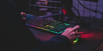 A person is playing a computer game, seen from the side. They are using a colorful RGB backlit keyboard and mouse, with a gaming monitor showing the game in the background. The dimly lit room adds to the gaming atmosphere. | FintechZoom