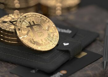 Close-up image of several gold-colored Bitcoin coins placed on a black leather Bitcoin wallet. Nearby, a black card with gold accents is partially visible alongside a pen. The background is slightly blurred, highlighting the coins and wallet. | FintechZoom