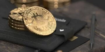Close-up image of several gold-colored Bitcoin coins placed on a black leather Bitcoin wallet. Nearby, a black card with gold accents is partially visible alongside a pen. The background is slightly blurred, highlighting the coins and wallet. | FintechZoom