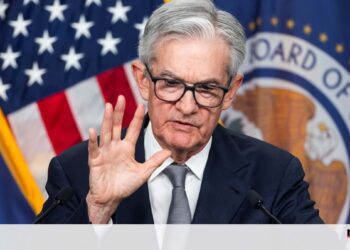 An older man with gray hair and glasses, dressed in a suit, speaks into a microphone while gesturing with his right hand. He stands in front of U.S. flags and a seal that reads "Board of Governors." The economic discussion is underscored by a logo in the bottom corner that reads "negocios. | FintechZoom