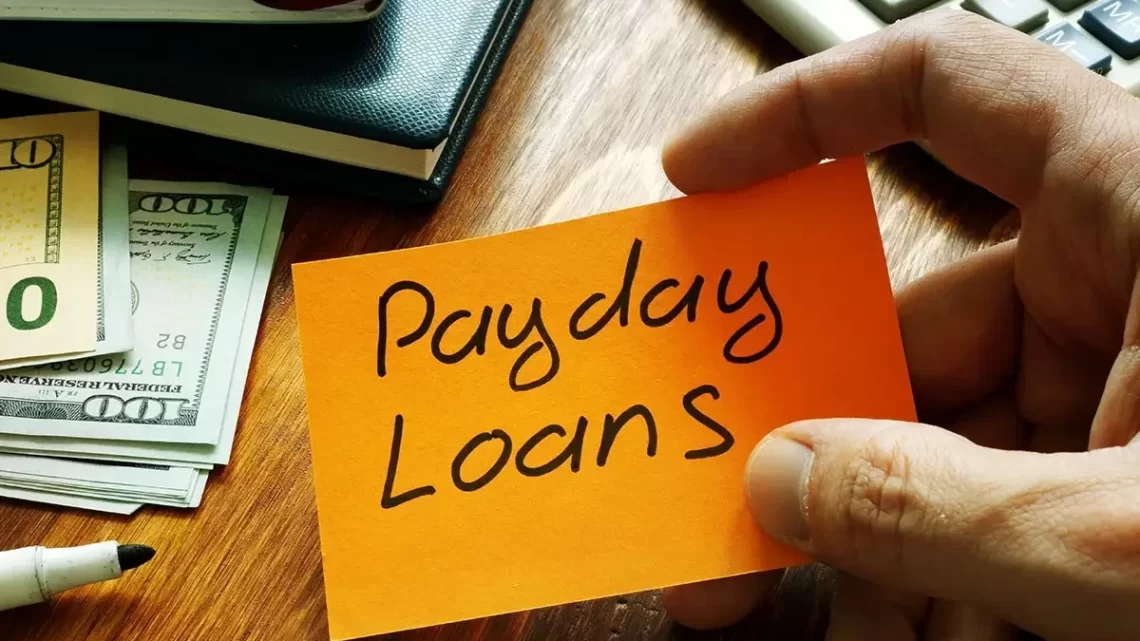 A hand holds an orange sticky note with "Payday Loans" written on it. In the background, a stack of 100 dollar bills, a pen, a notebook, and part of a calculator rest on a wooden surface. | FintechZoom