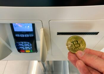 A person holding a physical representation of Bitcoin in front of a card reader and slot, possibly at a cryptocurrency payment gateway or digital currency exchange machine. | FintechZoom