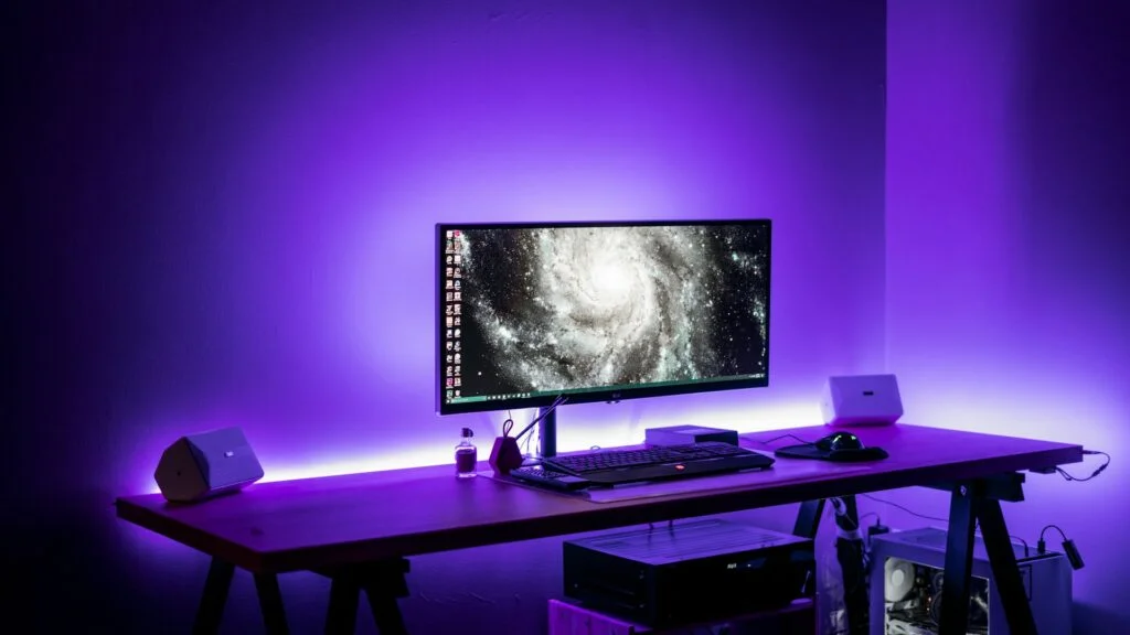 A modern desktop setup features a widescreen monitor displaying a galaxy image, flanked by two white speakers. The wooden desk is illuminated by purple LED lights, hosting a keyboard, mouse, and a few small accessories. A computer tower is placed underneath. | FintechZoom