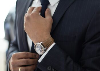 A close-up of a person adjusting their dark blue tie with one hand while wearing a dark pinstripe suit, exuding the precision of litigation in finance. They have a silver, white-faced wristwatch with a brown leather strap on their wrist. A wedding ring is visible on their other hand. | FintechZoom