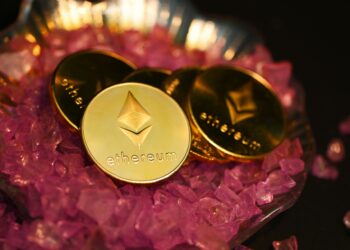 Three gold-colored Ethereum coins are arranged in a decorative dish filled with small, pink crystals, reminiscent of the glamor found in casinos. The Ethereum logo and the word "ethereum" are prominently displayed on each coin. The dark background makes the coins and crystals stand out elegantly. | FintechZoom