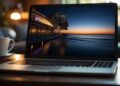 A laptop sits on a wooden table, displaying a serene beach scene at twilight. The screen shows calm waves and a pier with lit buildings, inviting thoughts of watching movies online. A white coffee cup and saucer are placed on the left side of the table. The background is softly blurred. | FintechZoom