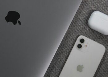 A space gray MacBook, a white iPhone with dual cameras, and a pair of white wireless earbuds in their charging case are placed on a textured gray fabric surface. The Apple logo is visible on both the MacBook and the iPhone, suggesting seamless integration with employee communication software. | FintechZoom