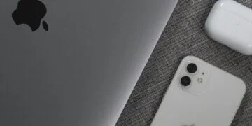 A space gray MacBook, a white iPhone with dual cameras, and a pair of white wireless earbuds in their charging case are placed on a textured gray fabric surface. The Apple logo is visible on both the MacBook and the iPhone, suggesting seamless integration with employee communication software. | FintechZoom