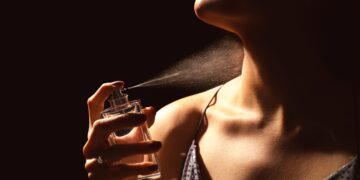 A woman applies the best perfume to their neck from a spray bottle. The image captures the fine mist mid-air, highlighting the person's neck and lower face against a dark background. They wear a sleeveless top with thin straps. | FintechZoom