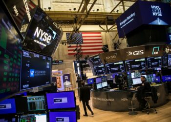 A bustling trading floor at the New York Stock Exchange features large digital screens displaying stock information, including the S&P 500. Traders work diligently at their stations under the glow of illuminated monitors, while an American flag is prominently displayed on the wall, encapsulating the high-energy environment. | FintechZoom