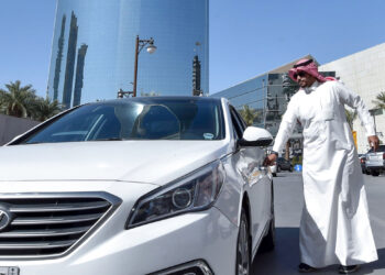 A man in traditional Middle Eastern attire stands next to a white car, about to open the driver's door. Behind him, modern glass buildings and palm trees are visible, indicating an urban setting with a mix of traditional and contemporary elements—a scene where the importance of car insurance in Saudi Arabia is evident. | FintechZoom