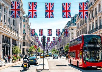A bustling street in London, vibrantly decorated with numerous Union Jack flags hanging overhead. Classic red double-decker buses, cars, pedestrians, and people on scooters traverse beneath a sunny, clear blue sky. Elegant historic buildings line both sides of the street, echoing the city's vibrant pulse akin to the FTSE 100 today. | FintechZoom