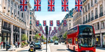 A bustling street in London, vibrantly decorated with numerous Union Jack flags hanging overhead. Classic red double-decker buses, cars, pedestrians, and people on scooters traverse beneath a sunny, clear blue sky. Elegant historic buildings line both sides of the street, echoing the city's vibrant pulse akin to the FTSE 100 today. | FintechZoom