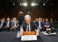 Jerome H. Powell sits at a table with a nameplate in front of him reading "The Honorable Jerome H. Powell" in a well-lit room, while screens displaying Nasdaq Today updates are visible in the background, suggesting a formal setting with numerous people, some seated and some standing. | FintechZoom