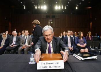 Jerome H. Powell sits at a table with a nameplate in front of him reading "The Honorable Jerome H. Powell" in a well-lit room, while screens displaying Nasdaq Today updates are visible in the background, suggesting a formal setting with numerous people, some seated and some standing. | FintechZoom