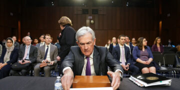 Jerome H. Powell sits at a table with a nameplate in front of him reading "The Honorable Jerome H. Powell" in a well-lit room, while screens displaying Nasdaq Today updates are visible in the background, suggesting a formal setting with numerous people, some seated and some standing. | FintechZoom