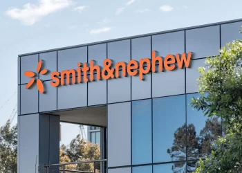 A modern office building with large reflective windows bears the "smith&nephew" logo in bright orange letters on its facade, hinting at their strong market presence as FTSE 100 rises 0.7% with Smith & Nephew leading gains. Trees and a clear blue sky are visible in the background. | FintechZoom