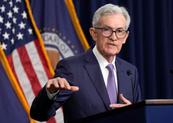 Jerome Powell , wearing a navy suit and purple tie, speaks at a podium with microphones. Behind him are the American flag and an official dark blue emblematic flag, indicating a formal or governmental setting. Jerome Powell discusses job openings | FintechZoom