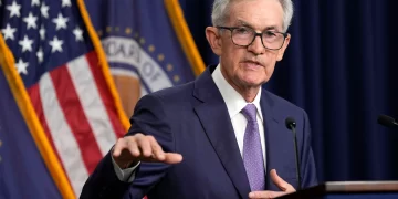 Jerome Powell , wearing a navy suit and purple tie, speaks at a podium with microphones. Behind him are the American flag and an official dark blue emblematic flag, indicating a formal or governmental setting. Jerome Powell discusses job openings | FintechZoom