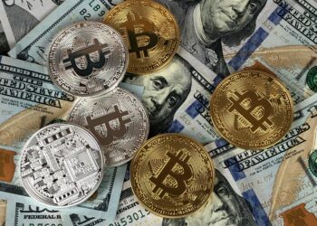 A mix of gold and silver physical Bitcoin tokens are placed on top of various denominations of U.S. dollar bills, including several visible $100 bills featuring Benjamin Franklin's portrait, symbolizing the intersection of traditional currency with cryptocurrencies. | FintechZoom