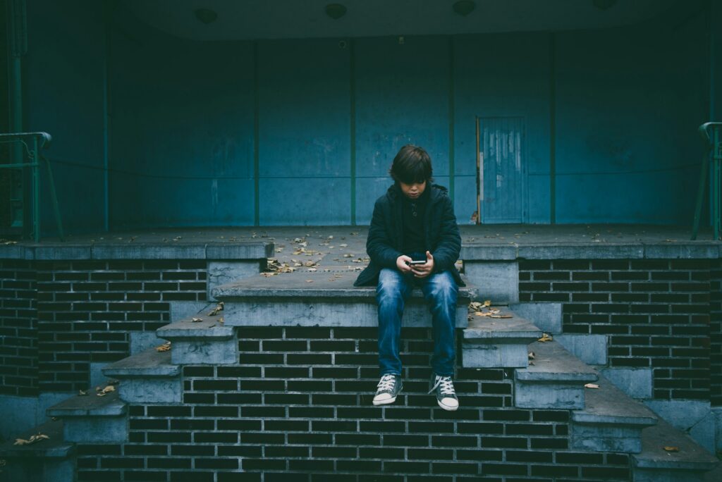 A child wearing a dark jacket and jeans sits alone on a set of concrete steps with a phone in hand, seemingly finding some comfort in the device. The background features a closed, gray building. Fallen leaves are scattered around. Despite the moody and somber atmosphere, there’s an underlying sense of safety. | FintechZoom