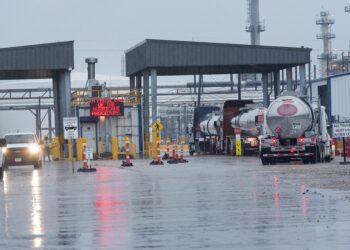 A queue of tanker trucks waits at an industrial facility's entrance on a rainy day. Vehicles pass through a covered gate with an electronic sign displaying "Hurricane Procedures." Oil prices today could be seen influencing the urgency, as the wet road reflects the scene against chimneys and pipes. | FintechZoom