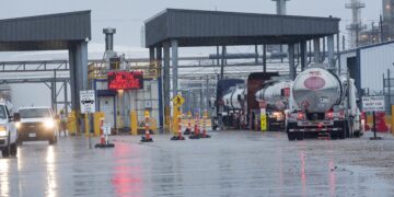 A queue of tanker trucks waits at an industrial facility's entrance on a rainy day. Vehicles pass through a covered gate with an electronic sign displaying "Hurricane Procedures." Oil prices today could be seen influencing the urgency, as the wet road reflects the scene against chimneys and pipes. | FintechZoom