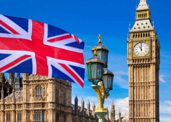 The image shows the British flag waving in front of the iconic Big Ben clock tower and the Houses of Parliament in London, under a clear blue sky. A decorative street lamp adds to the scene, symbolizing stability much like the FTSE 100 does for investors. | FintechZoom