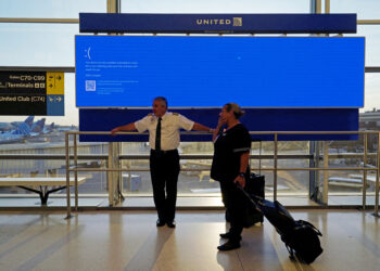 Two people in airport uniforms, one in a pilot's attire, stand before a large display screen showing a blue error message. Behind them, large windows reveal airplanes and airport facilities. Airline logos are visible in the background as the scene hints at the chaos caused by Microsoft's outage. | FintechZoom
