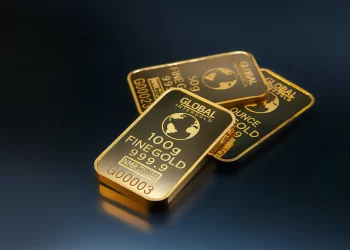 Three gold bars are stacked on a dark surface. The top bar is marked "GLOBAL INTERGOLD" and "100 g FINE GOLD 999.9" with a globe design. The other two bars display similar markings, including "1 OUNCE" and "INMR". These gleaming treasures, echoing the high gold price today, shine brightly reflecting the light. | FintechZoom