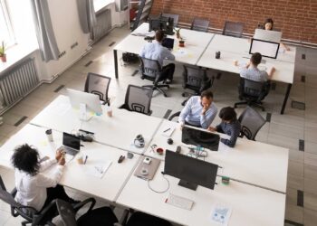 A modern open office space with six people working at their desks, embracing a desk-sharing culture. The office features white desks and grey chairs, multiple computers, and various office supplies. Large windows, brick walls, and a bright, clean atmosphere complete the setting. | FintechZoom