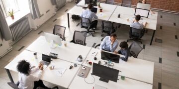 A modern open office space with six people working at their desks, embracing a desk-sharing culture. The office features white desks and grey chairs, multiple computers, and various office supplies. Large windows, brick walls, and a bright, clean atmosphere complete the setting. | FintechZoom