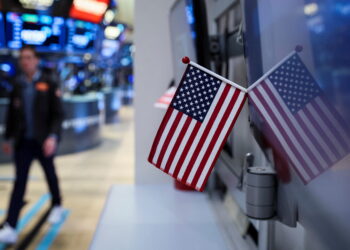 Two small American flags are mounted next to a computer screen, reflecting on its surface. In the background, a person walks through a busy stock exchange floor with digital screens and stock tickers displaying S&P 500 and Nasdaq 100 financial information. | FintechZoom