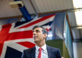 Rishi Sunak in a black suit with a white shirt and red tie is speaking in a room with a large UK flag hanging in the background. With a focused expression, he discusses the FTSE 100 as the ceiling and lights of the room are visible above him. | FintechZoom