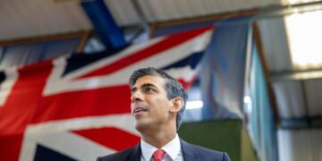 Rishi Sunak in a black suit with a white shirt and red tie is speaking in a room with a large UK flag hanging in the background. With a focused expression, he discusses the FTSE 100 as the ceiling and lights of the room are visible above him. | FintechZoom