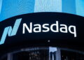 Brightly lit Nasdaq Today sign displayed on a large digital screen, with the Nasdaq logo consisting of a stylized letter "N" in shades of blue and the word "Nasdaq" in white text. The background features a modern building facade with reflective glass windows. | FintechZoom