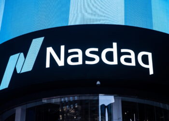 Brightly lit Nasdaq Today sign displayed on a large digital screen, with the Nasdaq logo consisting of a stylized letter "N" in shades of blue and the word "Nasdaq" in white text. The background features a modern building facade with reflective glass windows. | FintechZoom
