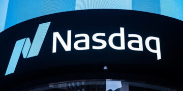 Brightly lit Nasdaq Today sign displayed on a large digital screen, with the Nasdaq logo consisting of a stylized letter "N" in shades of blue and the word "Nasdaq" in white text. The background features a modern building facade with reflective glass windows. | FintechZoom