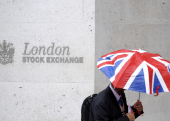 A person holding a Union Jack umbrella stands in front of a wall with the London Stock Exchange name and logo engraved on it. The individual, dressed in formal attire and carrying a backpack, appears to be checking the FTSE 100 today live updates. | FintechZoom