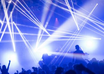 A vibrant nightclub scene with bright, colorful lights illuminating the stage and streaming in various directions. A DJ stands at the console, while a crowd of enthusiastic attendees raise their hands and dance energetically in the foreground. | FintechZoom