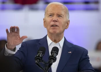 Biden, is speaking at a podium with two microphones. He has white hair and gestures with one hand raised. The background is blurred, featuring some people and a red, white, and blue flag partially visible. | FintechZoom