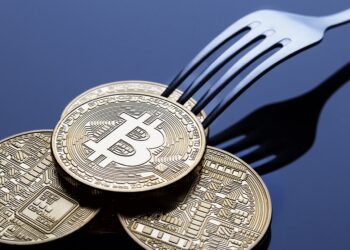 A shiny silver fork attempting to pick up three gold-colored physical Bitcoin tokens on a reflective dark surface, reminiscent of a bitcoin fork. The tokens display intricate circuit-like patterns and the prominent Bitcoin logo in the center. | FintechZoom