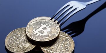 A shiny silver fork attempting to pick up three gold-colored physical Bitcoin tokens on a reflective dark surface, reminiscent of a bitcoin fork. The tokens display intricate circuit-like patterns and the prominent Bitcoin logo in the center. | FintechZoom