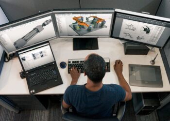 A person is working at a desk with a laptop and three large monitors displaying detailed technical drawings and 3D models, likely engineering designs created using CAD software. The individual is using a keyboard, mouse, and writing on a graphics tablet in an office cubicle. | FintechZoom